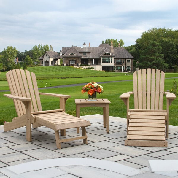 Two wooden Adirondack chairs with slatted backs and built-in ottomans are placed on a patio, accompanied by a small matching side table with a flower arrangement, overlooking a green lawn and a large house in the background.