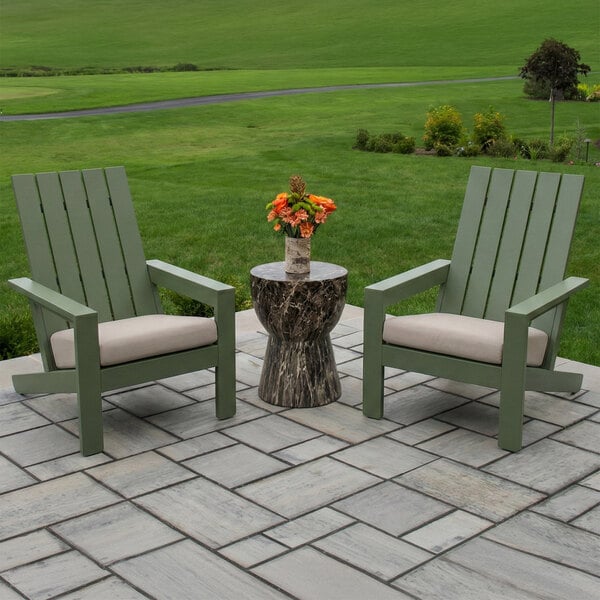 Two green Adirondack chairs with beige cushions are placed on a stone patio, with a small round table holding a vase of orange flowers between them, set against a grassy landscape.