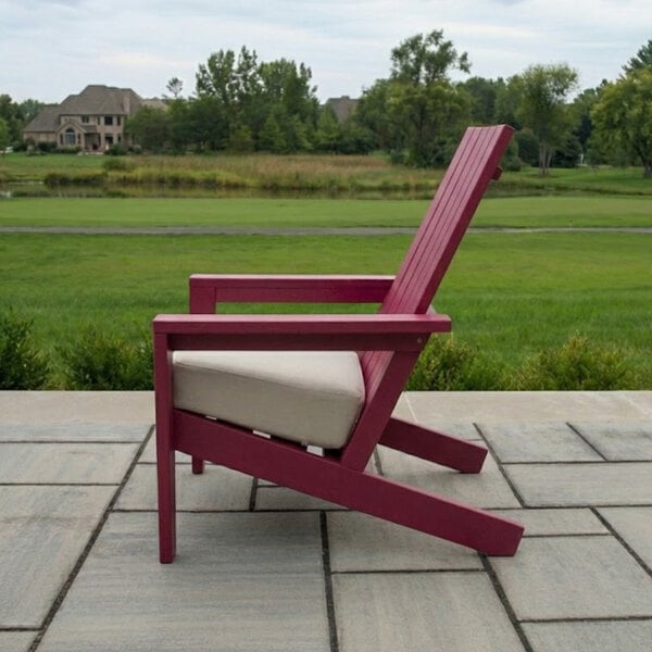 A side view of a pink Adirondack-style chair with a light-colored cushion, placed on a stone patio with a grassy landscape and trees in the background.