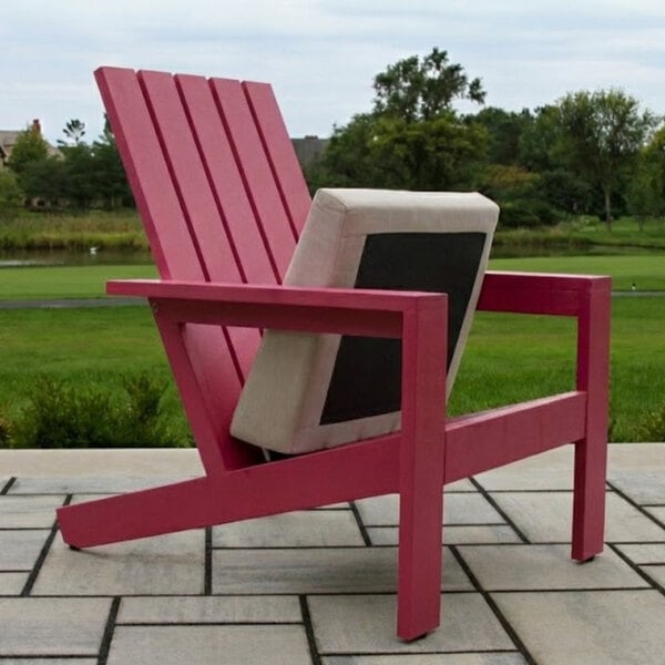 A pink Adirondack-style chair with a beige and black cushion placed on a patio with a grassy landscape in the background.