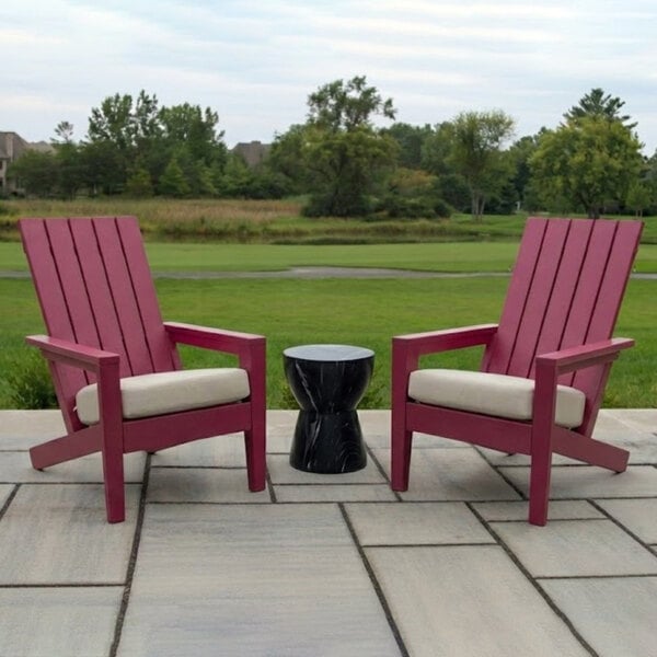 Two pink Adirondack-style chairs with white cushions are placed on a patio with a small black side table between them, overlooking a grassy landscape with trees in the background.