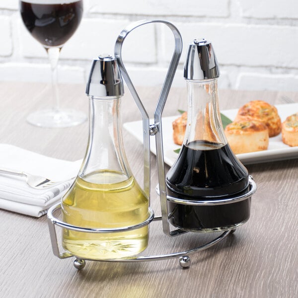 Two glass cruets, one filled with a light yellow liquid and the other with a dark liquid, are held in a chrome-plated holder on a wooden table.