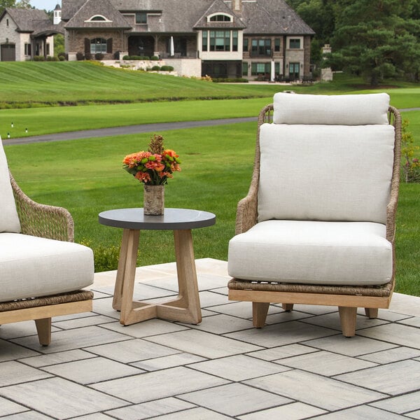 A small round table with a dark top and light wooden legs is placed outdoors between two cushioned chairs, with a vase of flowers on top.