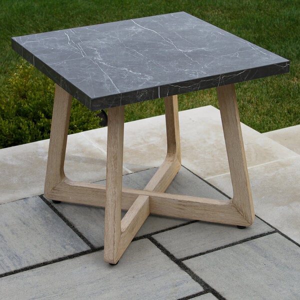 A square table with a dark, marble-like top and light wood legs, placed outdoors on a stone patio.