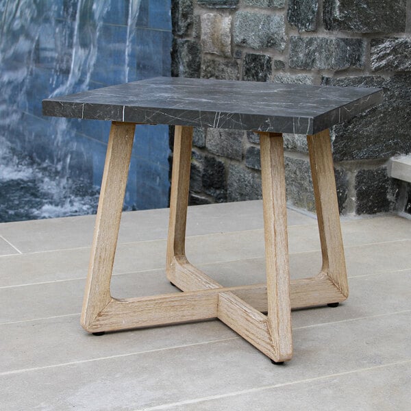 A square table with a dark stone-like top and light wood legs, placed outdoors on a tiled surface near a stone wall and water feature.