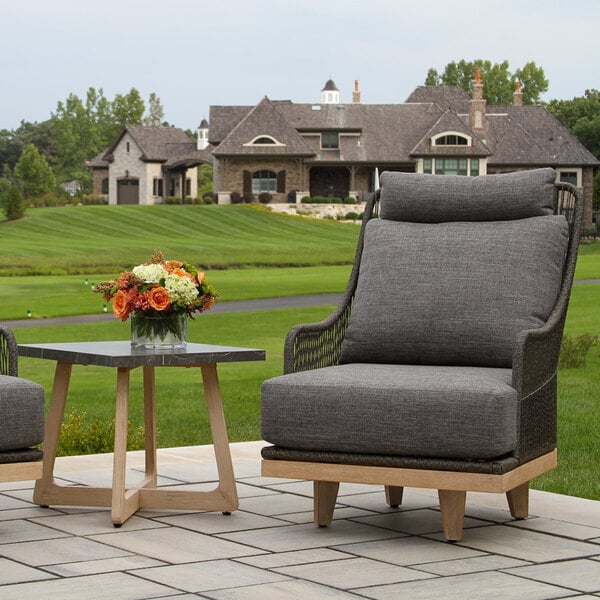 A small square table with a dark stone-like top and light wooden legs is placed outdoors next to a cushioned chair, with a flower arrangement on top.