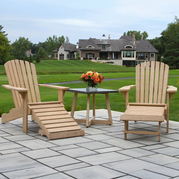 Two wooden Adirondack chairs with matching ottomans and a square accent table topped with a vase of flowers are arranged on a stone patio, with a large house and green lawn in the background.