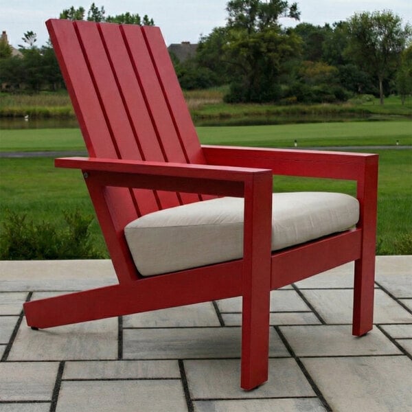 A red Adirondack-style chair with a slatted back and a light-colored cushion, placed on a patio with a grassy landscape in the background.