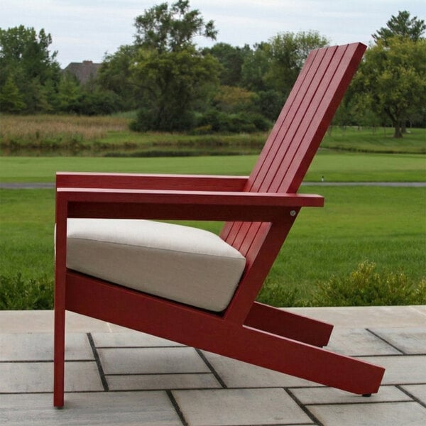 A red Adirondack-style chair with a beige cushion is placed on a patio with a grassy landscape in the background.