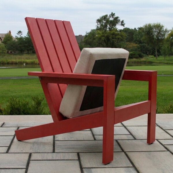 A red Adirondack-style chair with a beige and black cushion is placed on a tiled patio with a grassy landscape in the background.