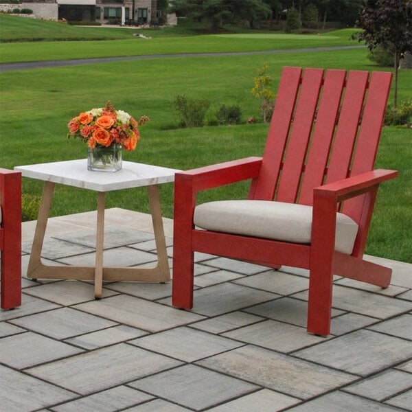 A red Adirondack chair with a light-colored cushion is placed on a stone patio next to a small table with a vase of orange flowers, with a grassy lawn in the background.