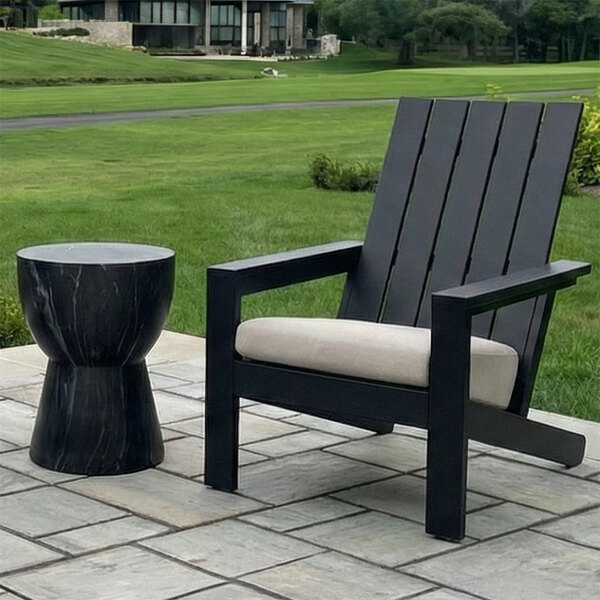 A black slatted outdoor chair with a light-colored cushion is placed next to a small, round, black side table on a stone patio with a grassy lawn in the background.
