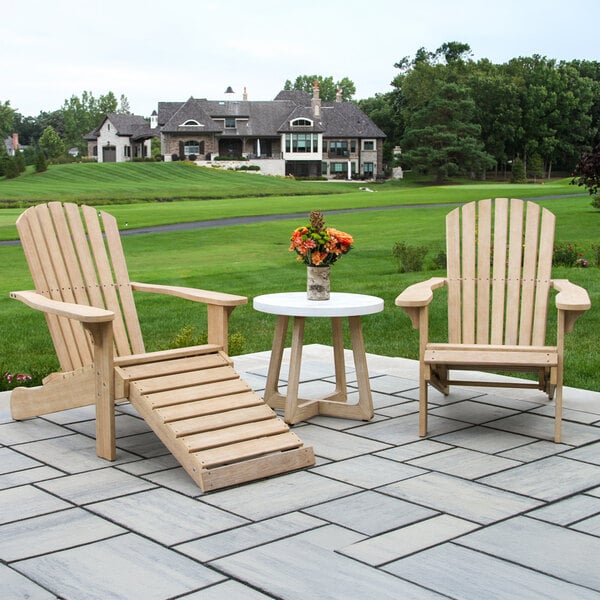 Two wooden Adirondack chairs with a matching ottoman and a small round table topped with a flower vase are arranged on a stone patio overlooking a green lawn and large house in the background.
