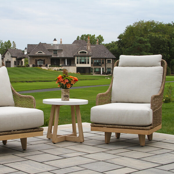 A small round table with a white top and wooden legs is placed between two cushioned outdoor chairs on a patio, with a vase of orange flowers on the table and a large house and green lawn in the background.