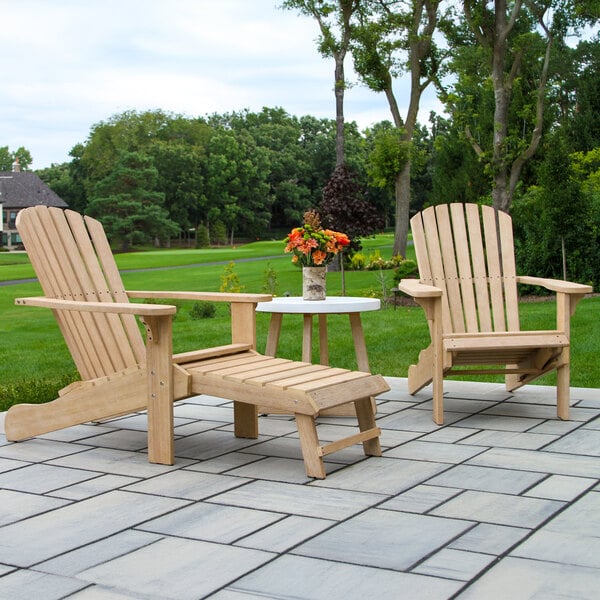 Two wooden Adirondack chairs with a matching footrest and a small round white table topped with a vase of flowers are arranged on a stone patio in a green outdoor setting.