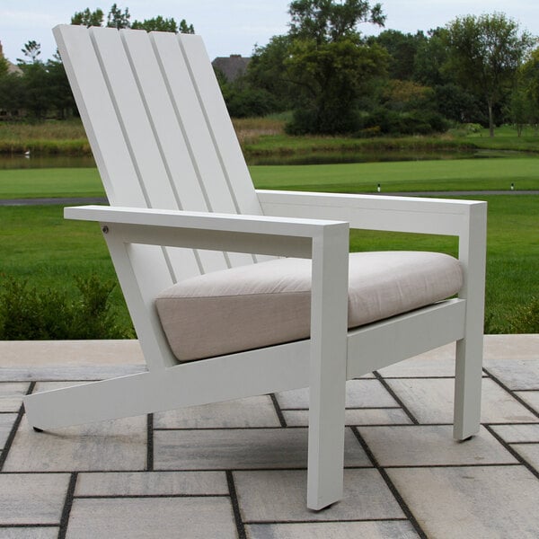 A white outdoor chair with a slatted backrest, straight armrests, and a light-colored seat cushion, placed on a tiled patio with a grassy landscape in the background.