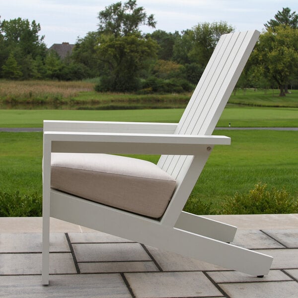A white slatted outdoor chair with a beige cushion, positioned on a stone patio with a grassy landscape in the background.