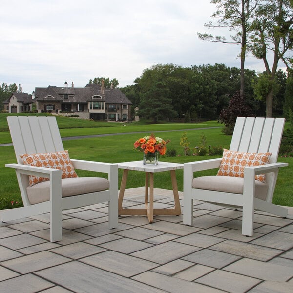 Two white Adirondack chairs with beige cushions and patterned orange pillows are positioned on a stone patio with a small wooden table holding a vase of orange flowers between them, overlooking a green lawn and large house in the background.