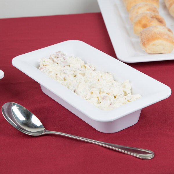 A white food pan filled with food next to a spoon on a red tablecloth.