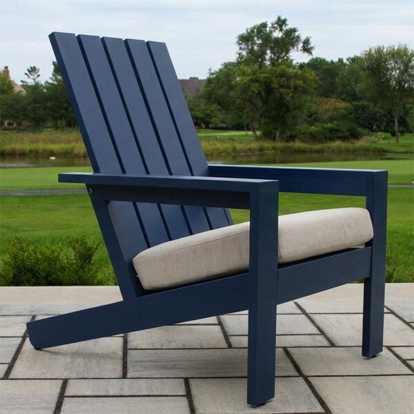 A blue Adirondack-style chair with a beige cushion is placed on a tiled patio with greenery and trees in the background.