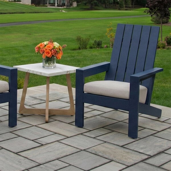 A blue Adirondack-style chair with a light-colored cushion is placed on a stone patio next to a small table with a vase of orange flowers, with a grassy lawn in the background.