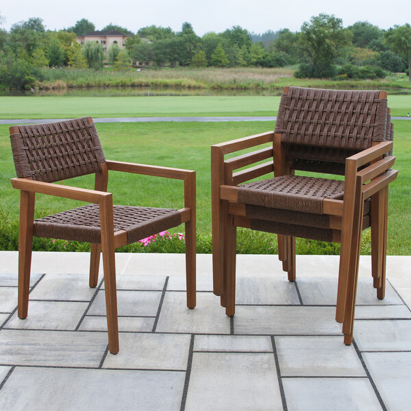 Two brown woven wicker and wood chairs are shown on a patio, with one chair standing alone and three stacked together, set against a green outdoor landscape.