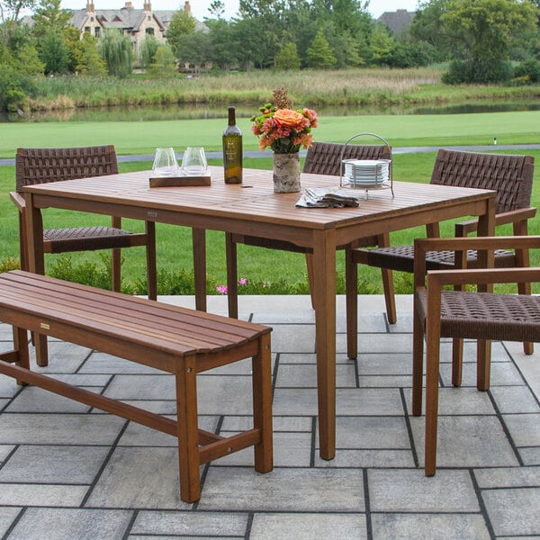 A wooden outdoor dining set with a rectangular table, matching bench, and woven wicker chairs, set on a patio with a scenic background.