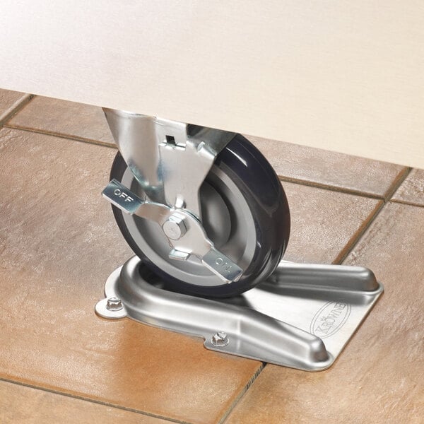 A metal caster wheel is positioned in a stainless steel holder mounted on a tiled floor.