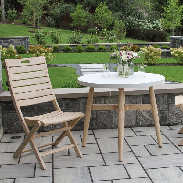 A round white table with wooden legs is paired with a wooden slatted chair on a stone patio, with glassware and a flower arrangement on the table, set in a landscaped garden.