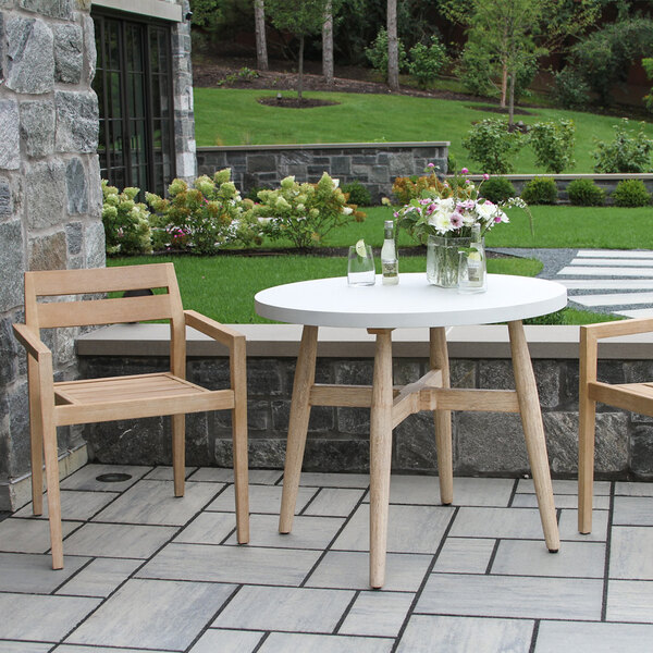 A round white table with wooden legs is placed outdoors on a tiled patio, accompanied by two wooden chairs, with a vase of flowers and bottles on the table.
