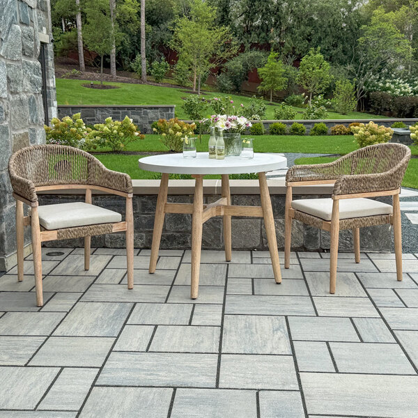 A round white table with wooden legs is paired with two wicker chairs with cushions, set on a tiled patio in a landscaped garden.