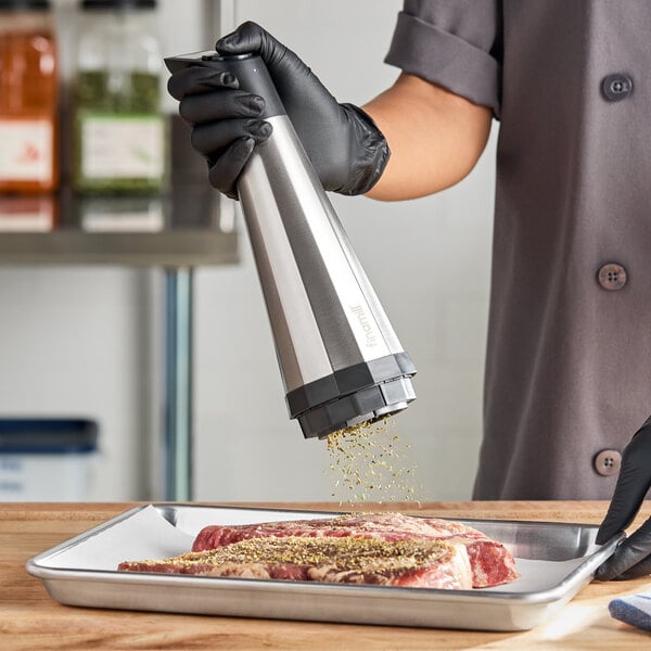 A person wearing black gloves is using a large, metallic electric spice grinder to season raw meat on a tray.