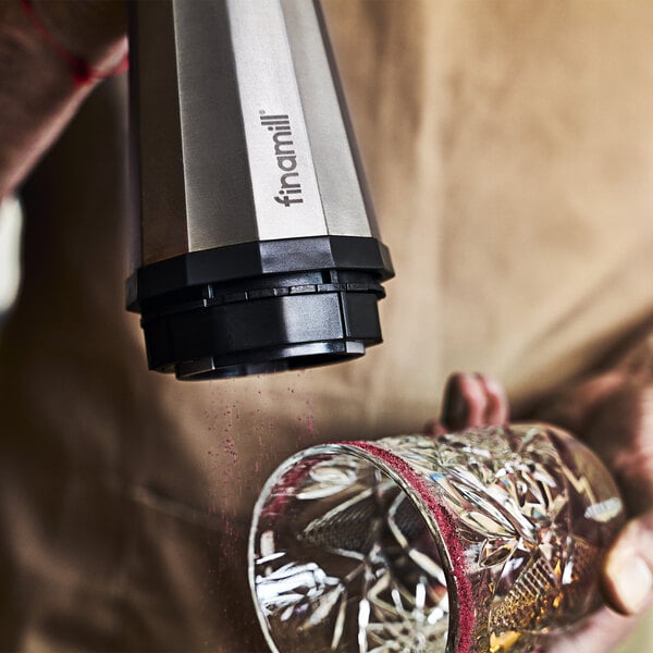 A close-up of a person using a metallic electric grinder to apply a reddish powder to the rim of a decorative glass.