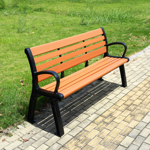 An orange slatted bench with black metal armrests and legs, placed on a paved walkway next to a grassy area.