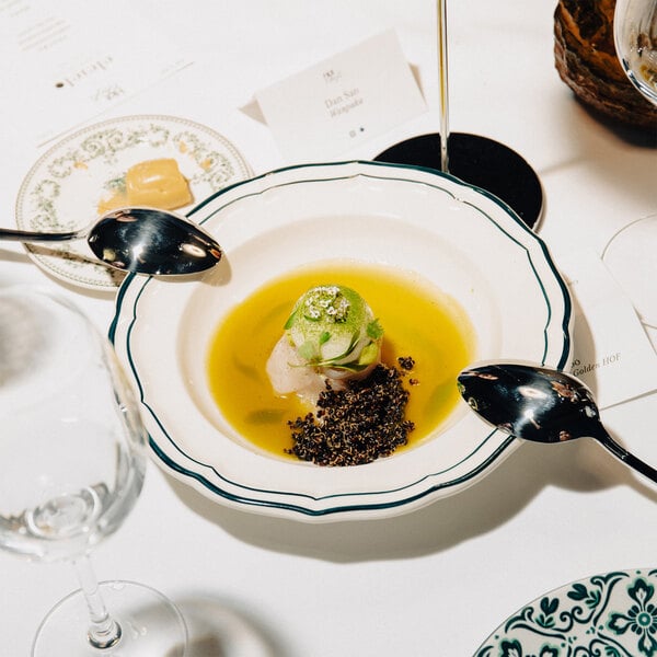 A white scalloped soup plate with a green rim, containing a gourmet dish with a yellow broth, a green-topped centerpiece, and a dark garnish, surrounded by two spoons.