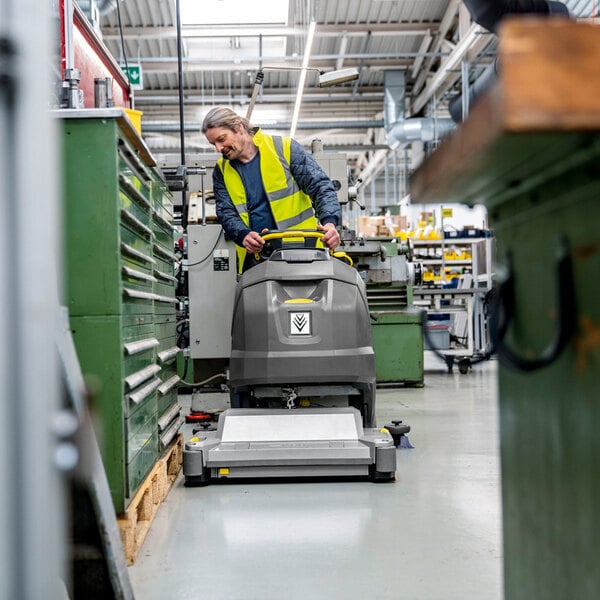 A person wearing a yellow safety vest operates a large, gray floor scrubber in an industrial workshop setting.