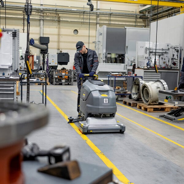 A person is operating a large, gray, walk-behind floor scrubber in an industrial workshop setting.