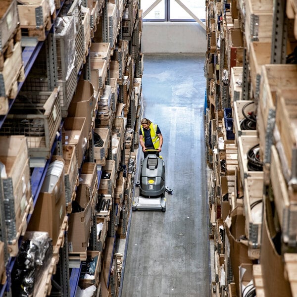 A person wearing a yellow safety vest is operating a floor scrubber in a warehouse aisle lined with shelves full of boxes and various items.