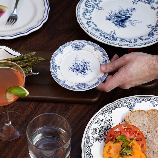 A hand is holding a small round white plate with blue floral patterns, surrounded by other patterned plates, food, and drinks on a wooden table.