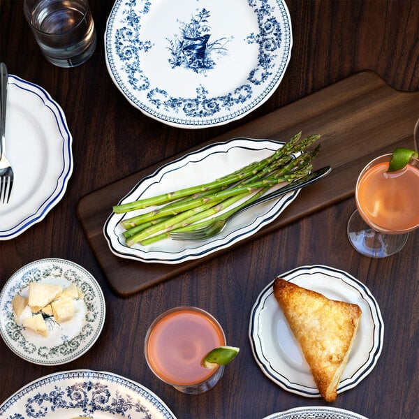 A table setting with various blue and white patterned plates, a scalloped dish holding asparagus, a triangular pastry, cheese, two pink cocktails with lime, and a glass of water on a dark wooden surface.