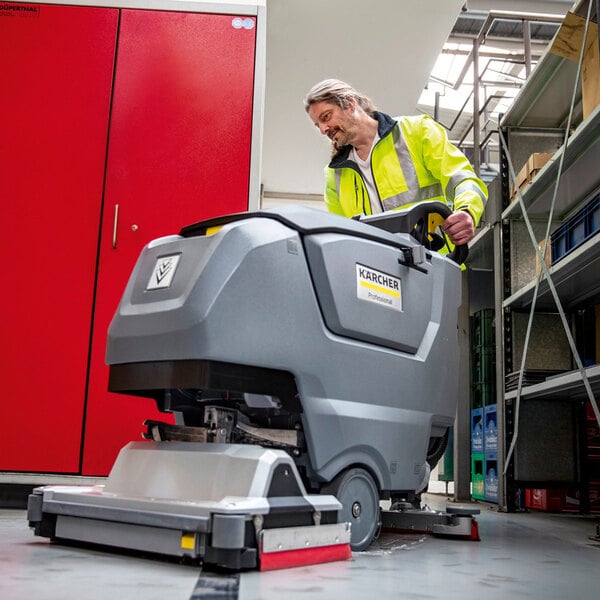A person in a high-visibility jacket operates a large, gray, walk-behind floor scrubber in an indoor industrial or commercial setting.