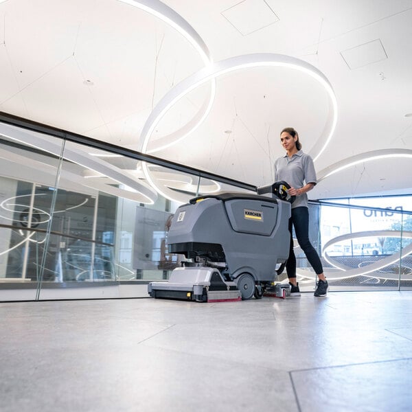 A person is operating a large, gray, walk-behind floor scrubber on a tiled floor in a modern, well-lit indoor space.
