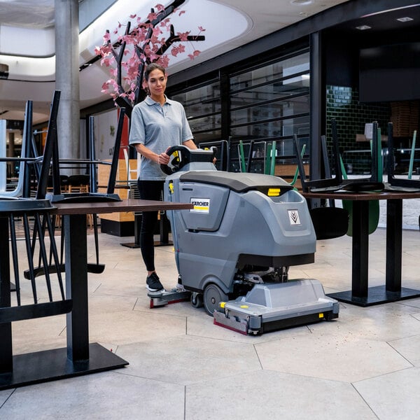A person is operating a large, gray, walk-behind floor scrubber in a commercial indoor space with tables and chairs.