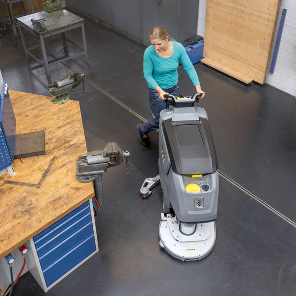 A person is operating a large, gray, walk-behind floor scrubber on a clean, dark floor in a workshop setting.