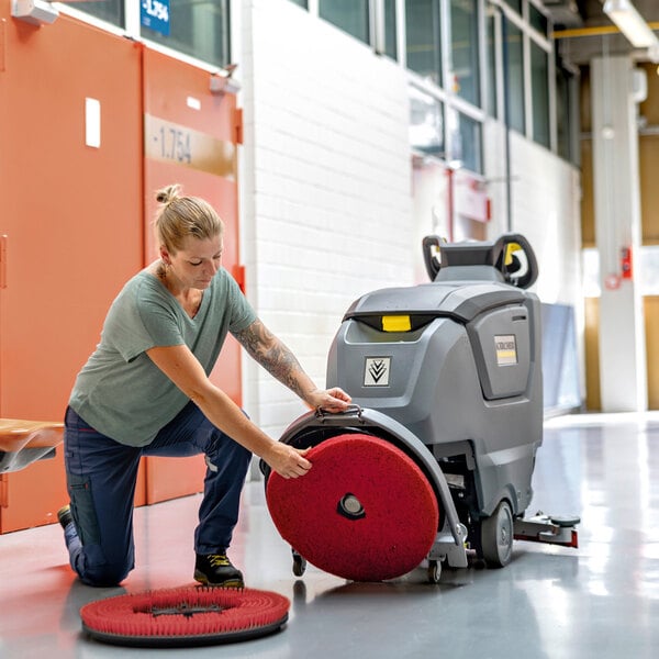 A person is kneeling on the floor, attaching a large red scrubbing pad to a gray walk-behind floor scrubber in a hallway.