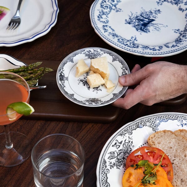 A hand is holding a small decorative plate with chunks of cheese, surrounded by other patterned plates, a cocktail, water glass, and food on a wooden table.