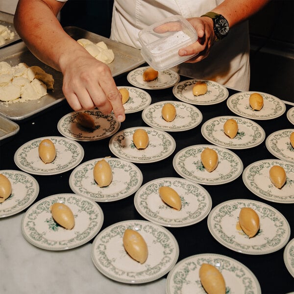 A person is placing small oval portions of food onto decorative white plates with green floral patterns, arranged in rows on a table.
