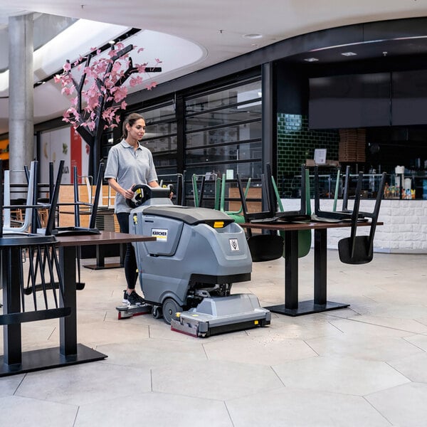 A person is operating a large, gray, walk-behind floor scrubber in a commercial space with tables and chairs.