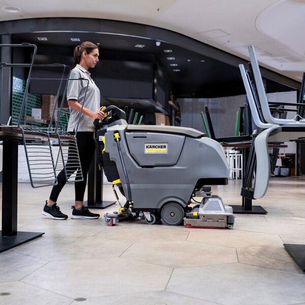 A person is operating a large, gray, walk-behind floor scrubber in a commercial indoor setting with chairs stacked on tables.