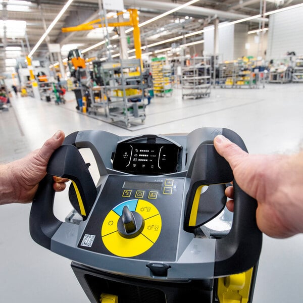 A person is operating a control panel with handles and a display screen in a large, well-lit industrial or manufacturing facility.